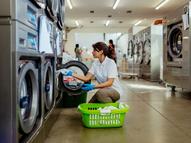 A worker in a commercial laundry facility handles clean linens with care, demonstrating the efficiency and professionalism of large-scale linen management operations. The scene includes industrial washing machines and organized storage systems.