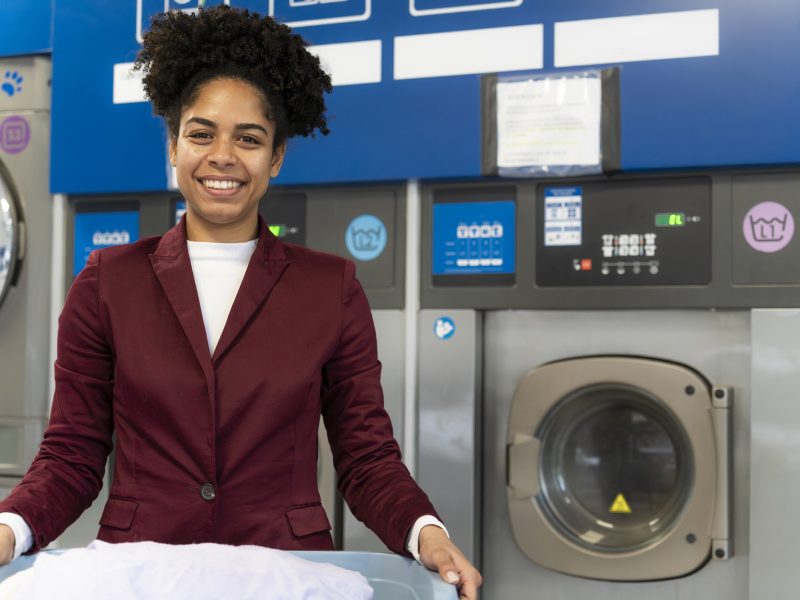 Young african american woman standing in self-service laundromat holding basket of white laundry and smiling