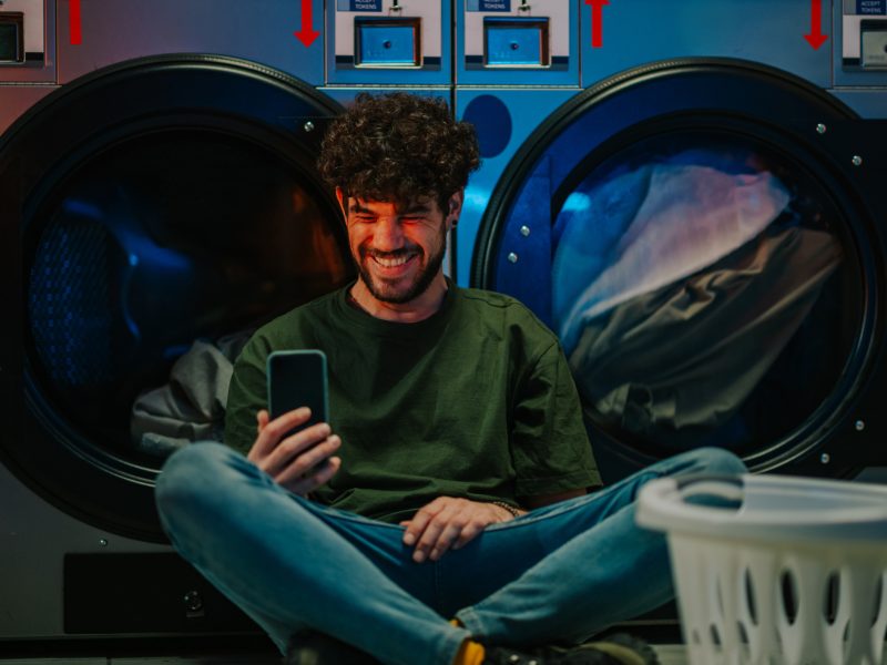 Cheerful young man using phone while waiting for laundry, sitting on laundromat floor